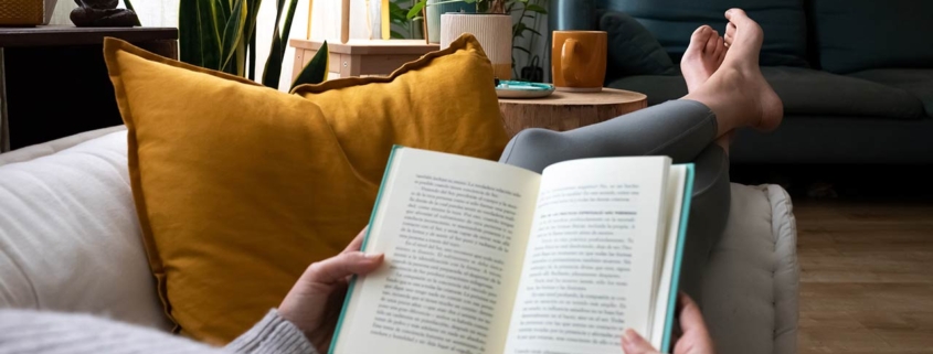 young woman relaxing at home reading a book lying on sofa