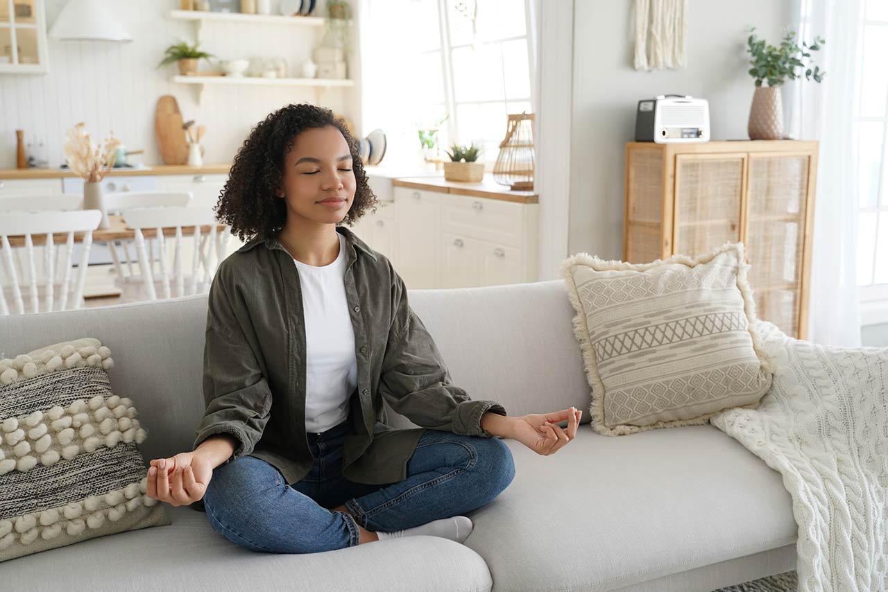 A serene Black woman meditates on a couch, eyes closed, in a sunlit, stylish living room