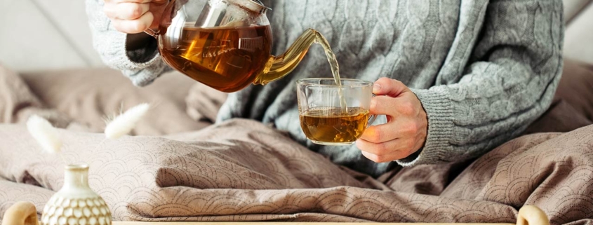 Woman dressed in knitted sweater pours hot tea from glass teapot into mug while sitting in bed