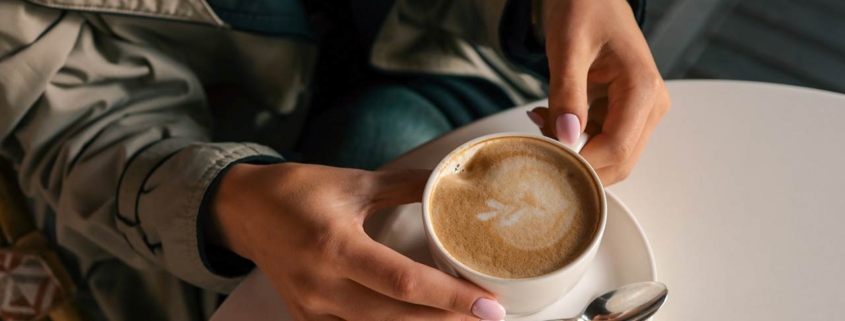 Close-up of woman's hands in trench coat gently holding a cup of cappuccino with latte art, seated at a white table